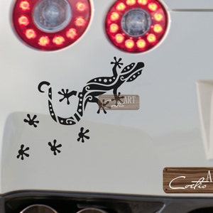 May include: Black gecko decal with detailed tribal patterns, applied to a white vehicle. The decal includes the word "ART" on a wooden sign. Additional black star-shaped decals are scattered around the gecko.