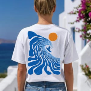 Op de afbeelding: Een wit t-shirt met korte mouwen en een grote blauwe afbeelding op de achterkant. Het ontwerp toont een gestileerde, golvende oceaan met het silhouet van een persoon op een surfplank en een oranje cirkel die de zon voorstelt.