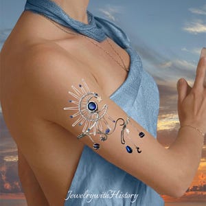 May include: A silver-toned arm cuff featuring a crescent moon and sunburst design with dark blue circular stones and small dangling star and moon charms. The metal wire structure wraps around the upper arm, accented with tiny blue beads.