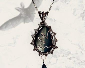 May include: A handcrafted pendant necklace featuring a dark, oval-shaped stone with a copper wire design. The pendant has a teardrop-shaped black crystal drop. The necklace is set against a marbled background with the text "JewelrywithHistory".