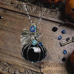 May include: A pendant necklace featuring a dark blue glittery stone shaped like a pumpkin, encased in silver-toned wire. The design includes a silver metal leaf, a small blue flower, a tiny butterfly, and a silver chain attached to the top.