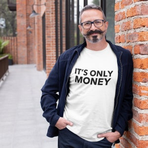 以下が含まれることがあります： 黒い文字で「IT'S ONLY MONEY」と書かれた白いTシャツを着た男性。青いジャケットと青いジーンズを着用しています。レンガの壁の前に立っています。