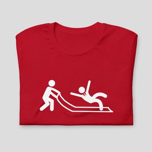 Puede incluir: Camiseta roja con un gráfico blanco de dos figuras de palo. Una figura de palo está empujando a la otra figura de palo hacia abajo por un tobogán.