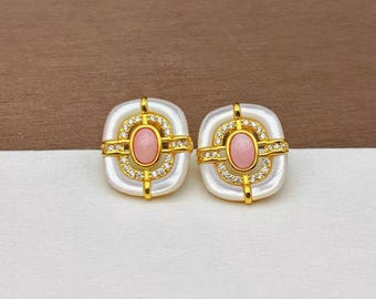May include: A pair of gold tone earrings with a white oval center and a pink oval stone in the center. The earrings are surrounded by clear rhinestones.