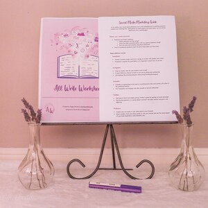 May include: A white booklet with the title "All Write Workshop" and a purple illustration of an open book with a cloud of ideas above it. The booklet is on a black metal stand with a purple marker on a white surface. Two clear glass vases with purple flowers are on either side of the stand.