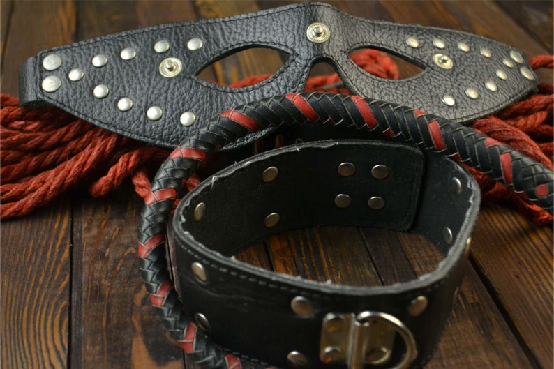 May include: A black leather mask with silver studs, a black leather choker with silver studs, and a black and red braided leather whip.