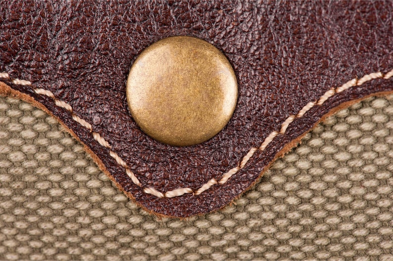 May include: Close-up of a brown leather patch with a stitched edge and a gold-toned metal button. The patch is sewn onto a beige canvas fabric.