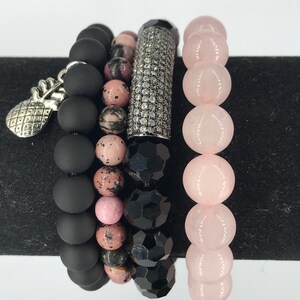 May include: A set of five bracelets featuring black matte beads, pink and black patterned beads, black faceted beads, and pink rose quartz beads. One bracelet has a silver pineapple charm.