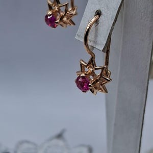 Puede incluir: Un par de pendientes dorados. Cada pendiente presenta un diseño de estrella con una gema roja redonda en el centro. Los pendientes se exhiben sobre una superficie blanca contra un fondo suave y borroso.