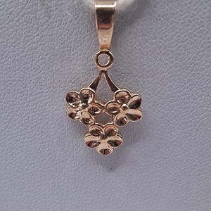Puede incluir: Un colgante de oro rosa con tres pequeñas flores de cinco pétalos dispuestas en un grupo triangular. El colgante está suspendido de una anilla sencilla de oro rosa pulido, sujeta a un cordel fino de color claro para su exhibición.