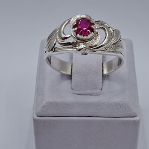 Könnte beinhalten: Ein silberner Ring mit einem leuchtend roten Edelstein. Das Ringdesign weist wirbelnde Muster und blattartige Details auf. Er wird auf einem kleinen, grauen Ständer vor weißem Hintergrund präsentiert. Das Design ist aufwendig.