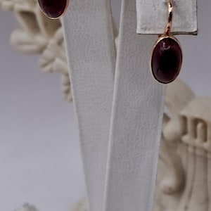 May include: Rose gold earrings with oval, dark red gemstones. The earrings are displayed on a white stand, showcasing their elegant design. The gemstones have a polished, reflective surface.
