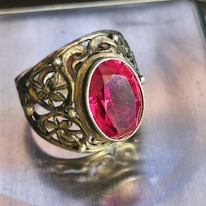 May include: A silver-toned ring featuring an oval-cut, vibrant pink gemstone set in a metal band with intricate, openwork floral filigree detailing. The band displays a weathered, textured finish across the metal surface surrounding the stone.