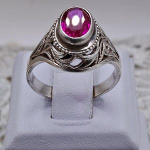 Può includere: Un anello d'argento con una grande gemma rosa vivace e rotonda. L'anello presenta un design a filigrana ornato attorno alla gemma e lungo la fascia. L'anello è esposto su una superficie bianca.