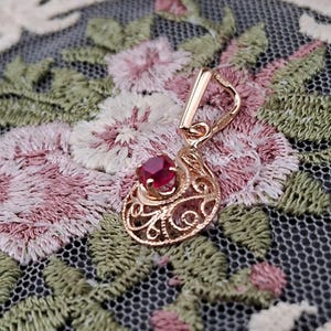 May include: A rose gold pendant featuring a circular filigree design with a single, small, round-cut, deep red gemstone set at the top. The pendant includes a polished rose gold bail at the top for attachment to a chain or necklace.