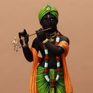 May include: A black statue of a figure playing a flute, adorned with a green turban, green and gold clothing, and a garland of white and red flowers. The figure has an orange shawl and is standing on a brown base.