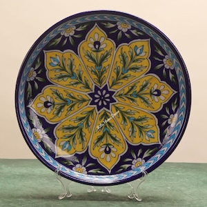 May include: A decorative ceramic plate with a floral design. The plate has a deep blue background with a large yellow flower in the centre, accented by green leaves and small white flowers. The rim has a blue and white pattern. The plate is round.