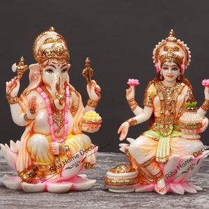 May include: A pair of decorative statues of Hindu deities, Ganesha and Lakshmi, sitting on lotus flower bases. Ganesha is depicted with an elephant head and four arms, holding a small axe, a lotus flower, a bowl of sweets, and a broken tusk. Lakshmi is depicted with four arms, holding a lotus flower, a pot of gold, and two other objects. Both statues are painted in gold, white, and pink.