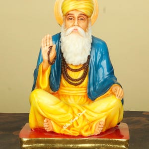 May include: A seated statue of a religious figure, wearing a yellow turban and robes, with a blue shawl. The figure has a white beard and is seated on a red and gold platform. A halo surrounds the head.