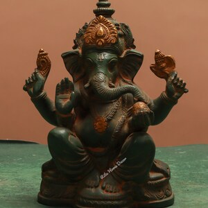 Puede incluir: Estatua sentada de la deidad hindú Ganesha, en una combinación de colores verde azulado y dorado. La estatua presenta detalles intrincados, incluyendo una corona, joyas y objetos en sus manos. La base es de color verde oscuro y el fondo es marrón sólido.
