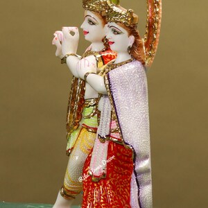 Puede incluir: Una estatua de m&aacute;rmol blanco de Radha Krishna, una pareja de deidades hind&uacute;es, con detalles intrincados y pintura colorida. La estatua est&aacute; de pie sobre una base de m&aacute;rmol blanco con acentos dorados.