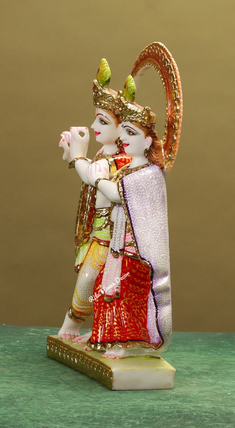 Puede incluir: Una estatua de m&aacute;rmol blanco de Radha Krishna, una pareja de deidades hind&uacute;es, con detalles intrincados y pintura colorida. La estatua est&aacute; de pie sobre una base de m&aacute;rmol blanco con acentos dorados.