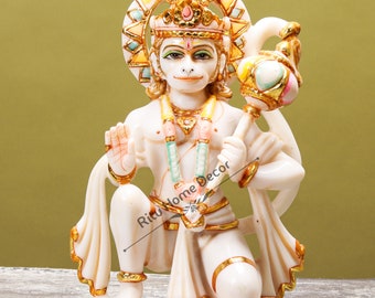 Może przedstawiać: Biały marmurowy posąg hinduskiego bóstwa Hanuman, klęczący z złotą koroną i trzymający laskę ze złotą kulą. Posąg jest ozdobiony złotą farbą i ma białą szatę opadającą na biodra.