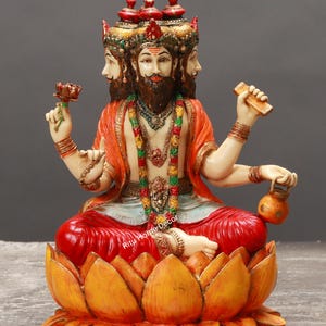 May include: A detailed statue of a deity with four faces, seated in a lotus position. The figure wears an orange robe, red pants, and ornate jewelry. The statue is holding a flower, a small container, and a wooden object. The base is a large, orange lotus flower.