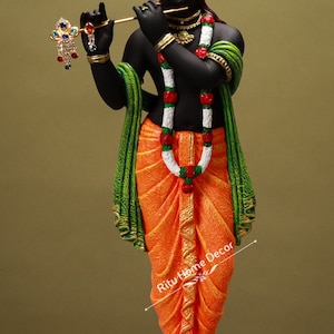 May include: A black statue of a deity, adorned with an orange turban and dhoti. The figure holds a flute and wears a garland of white and red flowers. Green and gold accents enhance the statue's details. The base is a reddish-brown color.
