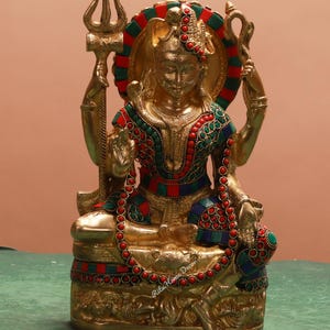 Puede incluir: Una estatua dorada de una deidad sentada, adornada con detalles rojos, verdes y azules. La estatua sostiene un tridente y una serpiente, con un halo con rayas rojas y verdes. La base es detallada y el diseño es ornamentado.