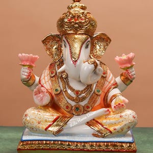 Peut inclure: Statue blanche et dorée de Ganesha, divinité hindoue, en position assise. La statue présente des détails dorés complexes sur la couronne, les bijoux et les vêtements. Ganesha tient des fleurs de lotus roses. La base est ornée de bordures dorées.