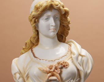 May include: White ceramic bust sculpture of a woman with a gold crown and a floral design on her chest. The bust is on a white pedestal. The text "Ritu Home Decor" is visible on the base.