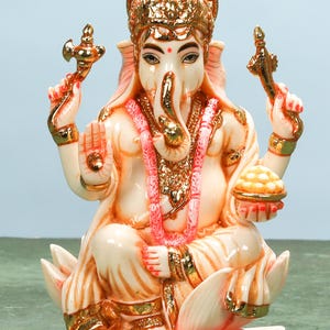 May include: A detailed statue of the Hindu deity Ganesha, seated on a lotus flower. The ivory-colored figure is adorned with gold accents, a pink garland, and a gold crown. Ganesha holds a sweet treat in one hand and a symbolic object in the other.
