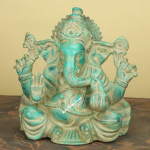 Puede incluir: Estatua turquesa y beige de la deidad hindú Ganesha, sentado en posición meditativa. La estatua presenta detalles intrincados, incluyendo una gran trompa, cabeza de elefante y múltiples brazos. La estatua tiene una apariencia envejecida y desgastada, con variaciones de textura y color visibles.