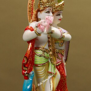 Puede incluir: Una estatua de m&aacute;rmol blanco de Radha Krishna, una pareja de deidades hind&uacute;es, con detalles intrincados y pintura colorida. La estatua presenta a Radha y Krishna de pie juntos, con Krishna tocando una flauta. La estatua est&aacute; decorada con acentos dorados y un tocado de plumas de pavo real.