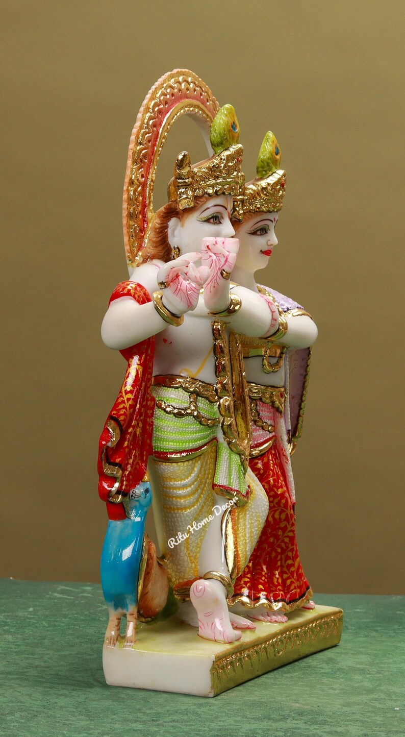Puede incluir: Una estatua de m&aacute;rmol blanco de Radha Krishna, una pareja de deidades hind&uacute;es, con detalles intrincados y pintura colorida. La estatua presenta a Radha y Krishna de pie juntos, con Krishna tocando una flauta. La estatua est&aacute; decorada con acentos dorados y un tocado de plumas de pavo real.