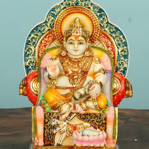 Może przedstawiać: Szczegółowa statua bóstwa siedzącego na tronie. Postać jest ozdobiona złotą biżuterią i koroną, z pogodnym wyrazem twarzy. Tron jest misternie zdobiony kolorowymi wzorami i złotymi akcentami, na jasnoniebieskim tle.