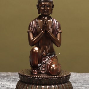 May include: A bronze statue of a kneeling figure with hands clasped in prayer. The figure is wearing a beaded necklace and a decorative belt. The statue is sitting on a lotus-shaped base.