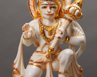 Może przedstawiać: Biała marmurowa statua hinduskiego bóstwa Hanuman, przedstawiająca go klęczącego z złotą laską w prawej ręce. Ma na sobie złotą koronę i biały materiał owinięty wokół ciała. Statua jest ozdobiona złotymi akcentami i stoi na białej marmurowej podstawie.