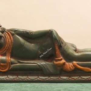 Puede incluir: Estatua reclinada de una deidad, cuerpo verde con detalles en naranja y marrón. La figura yace sobre una base rectangular con patrones tallados. La estatua tiene una expresión serena y está adornada con joyas detalladas. Mide aproximadamente 30 cm de largo.