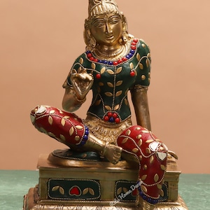 May include: A brass statue of a seated figure with detailed craftsmanship. The figure is dressed in green and red attire, embellished with decorative features. The statue rests on a decorative base, displaying a mix of colours and textures.