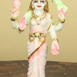 May include: A white and pink statue of a deity with multiple arms, adorned with gold accents. The figure wears a gold crown and jewelry, holding a bird and a decorative item. The base is white with gold trim. The statue is a religious or decorative item.