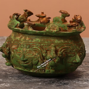 May include: Green ceramic pot with intricate carvings of faces and figures. The pot has a wide rim with decorative gold accents.