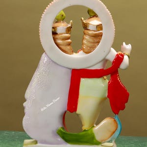 Puede incluir: Una figurita blanca, multicolor y con detalles dorados de una deidad hind&uacute;, Radha Krishna, de pie sobre una base blanca. La figurita sostiene una flauta y tiene una bufanda roja. La deidad est&aacute; representada con un halo y un marco circular alrededor de la cabeza. El texto "Ritu Home Decor" es visible en la figurita.