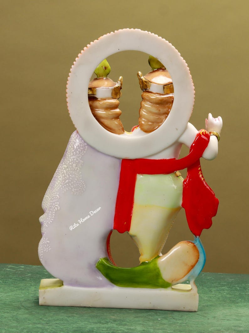 Puede incluir: Una figurita blanca, multicolor y con detalles dorados de una deidad hind&uacute;, Radha Krishna, de pie sobre una base blanca. La figurita sostiene una flauta y tiene una bufanda roja. La deidad est&aacute; representada con un halo y un marco circular alrededor de la cabeza. El texto "Ritu Home Decor" es visible en la figurita.