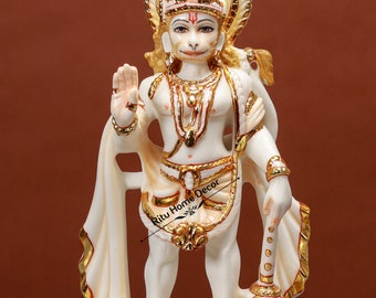 Może przedstawiać: Biała marmurowa statua hinduskiego bóstwa Hanuman, stojąca z złotą laską w prawej ręce. Ma na sobie biały dhoti i złotą koronę. Statua stoi na białej podstawie ze złotymi akcentami.