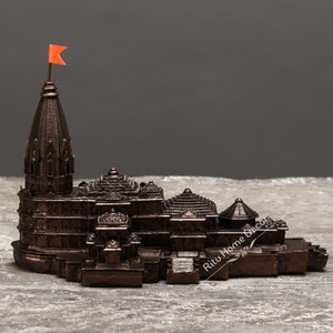May include: A small, detailed replica of a Hindu temple made of dark brown metal. The temple has a tall, pointed spire with a small orange flag on top. The temple is sitting on a grey surface.