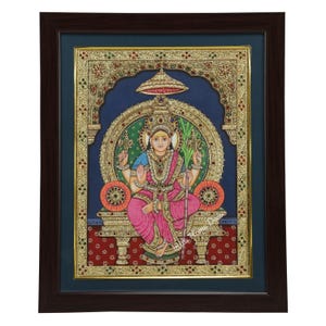 Puede incluir: Pintura enmarcada y ornamentada de una deidad sentada, con colores vibrantes y detalles intrincados. La figura viste una prenda rosa y está adornada con joyas de oro. El fondo es una mezcla de azul, verde y oro, con un marco marrón oscuro.