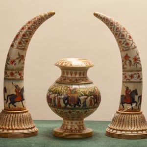 May include: Decorative ivory-colored sculptures. Two curved, horn-shaped pieces flank a central vase. The horn-shaped pieces feature floral patterns and figures on horseback. The vase has a bulbous shape with a detailed painted scene.