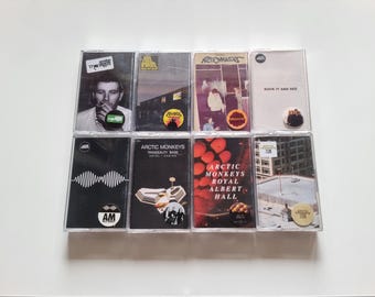 May include: A collection of eight Arctic Monkeys music cassette tapes arranged in two rows. The covers feature various album artworks including portraits, architectural scenes, and graphic designs, with some tapes displaying circular stickers.